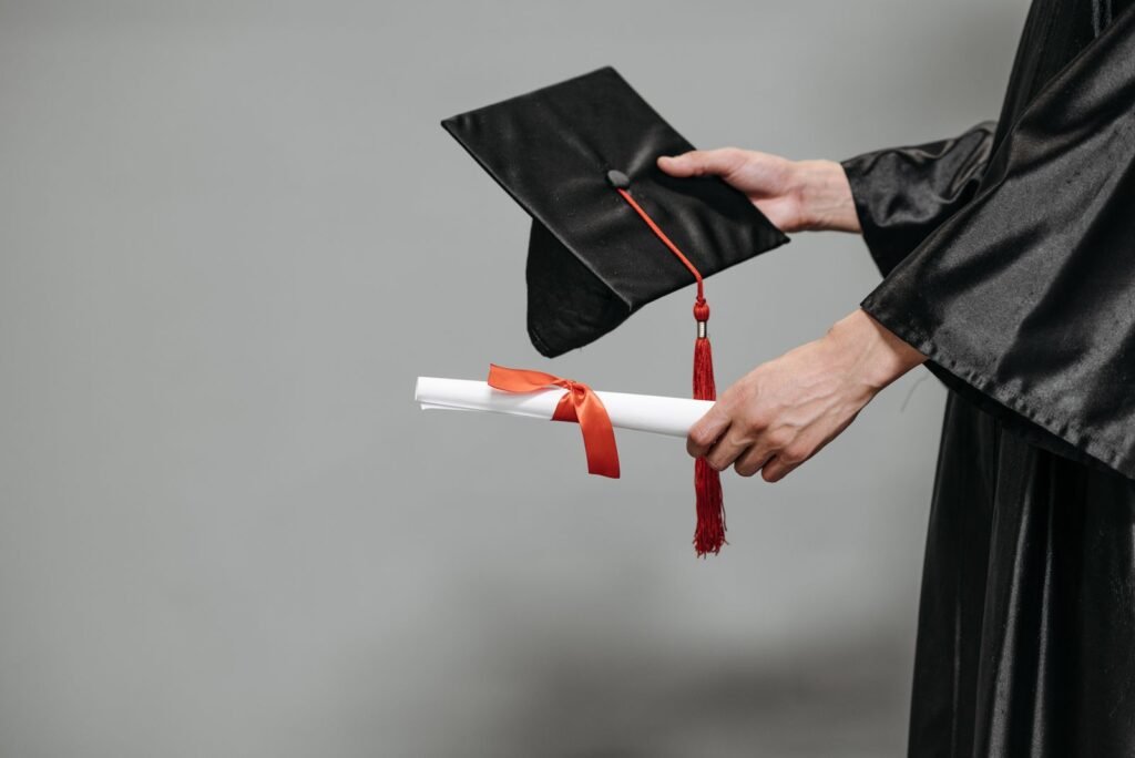Osoba u akademskoj odori drži crnu diplomsku kapu i uvijenu diplomu sa crvenom trakom.
